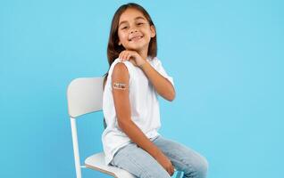 Vaccination Of Children. Happy Vaccinated Kid Girl Showing Arm With Adhesive Bandage After Vaccine Injection Sitting On Blue Studio Background. Kids And Covid-19 Prevention, Antiviral Immunization photo