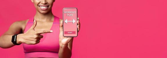 Cropped of smiling afro fitness lady pointing at workout application on mobile phone screen, red background, free space photo