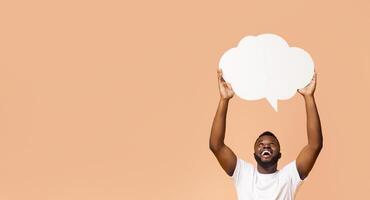 Happy Black man holding a blank speech bubble up above his head with his arms raised. He is looking up at the bubble and smiling broadly. The background is a simple peach color, copy space photo