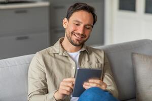 A man sits on a gray couch in a living room, smiling while using a tablet. He is wearing a beige button-down shirt and blue jeans. He is looking down at the tablet. photo