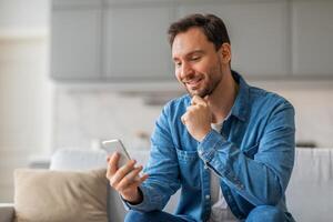 A man with a beard sits on a couch and looks at a smartphone with a smile on his face. He is wearing a blue button-up shirt and jeans. The background shows a modern home interior, copy space photo
