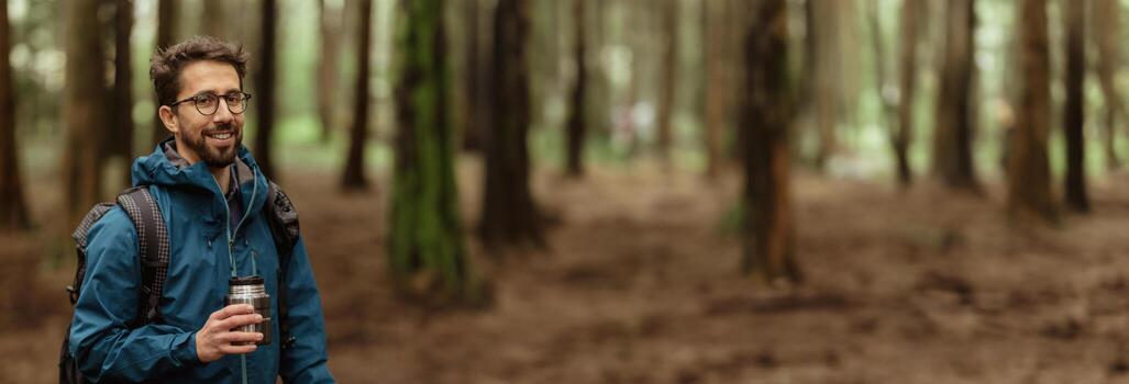A man with a backpack and a reusable coffee cup walks through a forest. The man is smiling and appears to be enjoying his hike. The forest is dense with trees, panorama with copy space photo