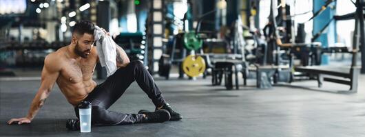 A muscular man sits on the gym floor, wiping sweat from his brow, after completing a strenuous workout. He holds a towel and a water bottle nearby photo
