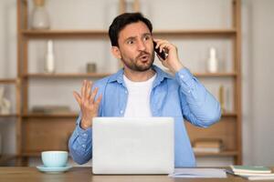 A man appears troubled or confused as he talks on his mobile phone with one hand gesturing, sitting in front of an open laptop, possibly working from home or an office photo