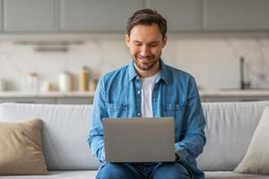 A man wearing a blue denim shirt and jeans sits on a white couch in a modern home, using a laptop computer. He is smiling and looking down at the screen, possibly working or browsing the internet. photo
