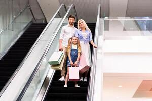 A happy family, consisting of a father, a mother, and a young girl, ride an escalator upward in a modern shopping mall. They are all smiling and holding shopping bags photo