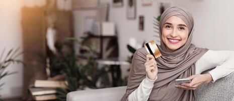 Online banking. Portrait of happy arabic woman in hijab with smartphone and credit card sitting on sofa at home, enjoying new app for payments photo