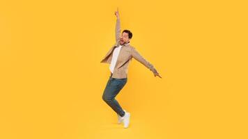 A man with a beard and a tan jacket jumps in the air with his right arm raised, pointing upward. He is wearing blue jeans and white sneakers and is facing the camera. He has a shocked expression photo