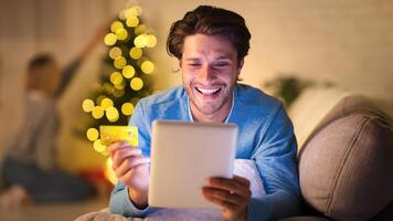 Handsome man shopping online on tablet on New Year's eve, woman decorating Christmas tree on background photo