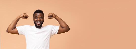 A happy, young, Black man with a short beard is flexing his muscles while standing in front of a beige background. He is wearing a white t-shirt and has a big smile on his face, copy space, panorama photo