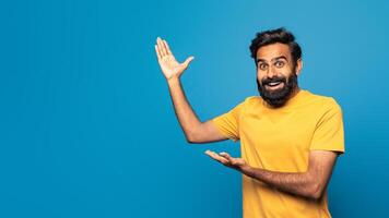 A cheerful Indian man wearing a yellow shirt smiles widely while enthusiastically gesturing with both hands in a studio against a vibrant blue background, copy space photo