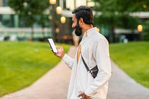 Indian man dressed in a light-colored shirt walks down a pathway in a vibrant urban park during the early evening. He is attentively looking at his smartphone, possibly checking messages or browsing. photo