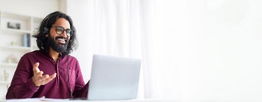 Online Coaching. Smiling Indian Guy Tutor Making Call With Laptop And Headset, Happy Young Eastern Man Sitting At Desk In Home Office, Talking And Gesturing At Web Camera, Copy Space photo