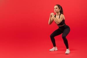 A focused young woman wearing a black sports outfit with white sneakers is engaged in a squatting exercise. Her hands are clenched together in front of her as she maintains a proper form, copy space photo