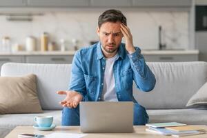 A man sits on a couch at home, looking frustrated and confused while using a laptop. He has one hand on his head and is gesturing with his other hand, as if he is trying to explain something. photo