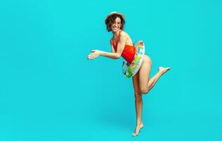 A happy black woman with curly hair is wearing a red swimsuit and a straw hat. She is holding an inflatable ring in one hand, and she is posing against a blue background. photo