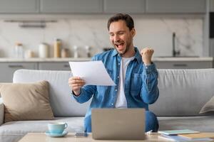 A man is sitting on a couch in a modern living room, holding a piece of paper and smiling widely. He has his right fist raised in the air in a gesture of celebration. He is likely reading good news photo