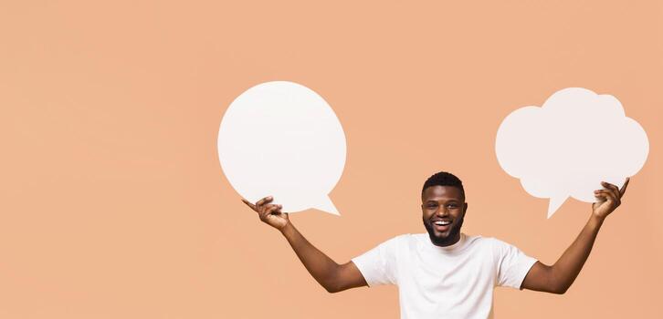 A happy man with a beard is holding up two blank speech bubbles, one round and one cloud-shaped, in front of a peach-colored background. He is looking directly at the camera with a wide smile. photo