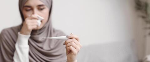 Sick islamic woman in hijab looking at thermometer, checking her temperature, suffering from seasonal flu at home, selective focus photo