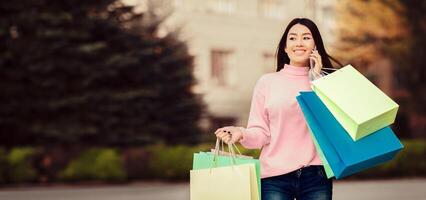 Asian woman is shown carrying multiple shopping bags while engaged in a phone conversation. She appears focused on her call while navigating through a crowded area with her purchases. photo