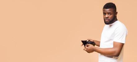A young African American man in a white shirt stands in front of a beige background, holding his empty wallet open and looking towards the camera with a concerned expression, copy space photo