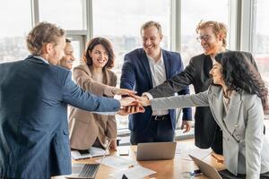 A diverse business team of six people stand around a table with laptops and paperwork in a modern office. They have their hands stacked in the center, smiling and celebrating a shared success. photo