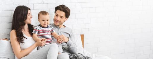 A joyful young couple sits on a bed, engaging with their cheerful baby. The parents smile at each other and the child, creating a warm atmosphere. Soft light fills the room, copy space photo