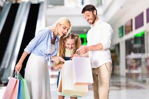 A young girl is standing between her parents, all three looking down at a pile of shopping bags. The girl is smiling, and the parents are smiling as they look at the contents of the bags. photo