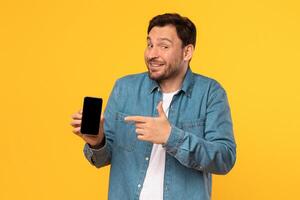 A man in a blue denim shirt stands in front of a yellow background. He is holding a smartphone in his left hand and pointing at the screen with his right hand. He has a happy expression on his face. photo