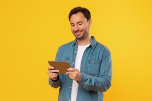 A man wearing a denim shirt stands in front of a bright yellow background, holding a tablet computer in his hands and looking down at the screen. He is smiling as he interacts with the tablet. photo