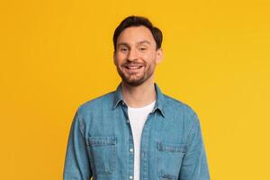A man wearing a blue denim shirt is standing in front of a solid yellow background. He is smiling at the camera with a friendly expression. photo