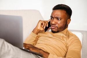 Computer Problem. Discontended Afro Guy Talking On Phone Frowning And Using Laptop Lying On Sofa At Home. Selective Focus photo