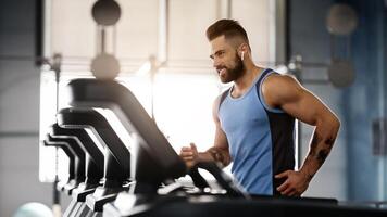 Jogging with pleasure. Young handsome man in modern wireless headphones smiling, running on treadmill at gym, free space photo