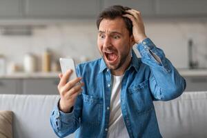 A man in a blue denim shirt is sitting on a couch in a kitchen. His mouth is open in shock, and he has his hand on his head. He is looking at a phone screen in his other hand. photo