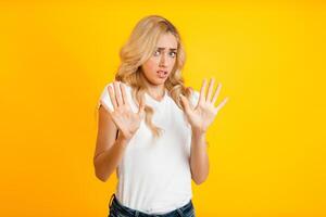 A woman with blonde hair stands in front of a yellow background, showing both hands in a stop gesture. She has a concerned expression on her face, trying to deter someone from approaching. photo