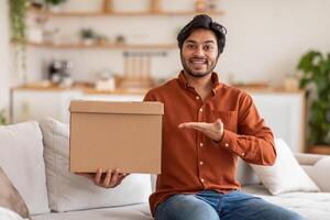 Arab man in an orange shirt sits on a couch, holding a cardboard box and smiling at the camera. One hand gestures towards the box, while the other holds the box in front of his waist. photo