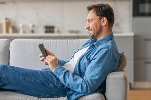 A man is sitting on a couch in a living room while using a smartphone. He is smiling as he looks at the phone. He is wearing jeans and a blue denim shirt. photo