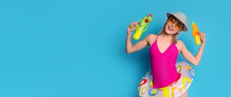 A young woman in a pink swimsuit, a sun hat, and sunglasses holds two water guns. She smiles at the camera with a playful expression. Her joyful demeanor suggests fun and excitement photo