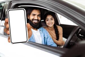 A bearded Indian man is extending his arm out of a car window, holding a smartphone with a blank screen towards the camera possibly for an interactive display or advertising purpose. photo