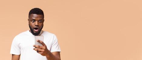 Black man with a shocked expression looks at his smartphone while standing in front of a beige background. His mouth is open and his eyes are wide, copy space photo