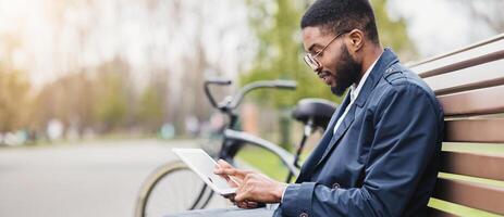 A professional black man is seated on a park bench, using a tablet with his bicycle next to him. The scene is peaceful and modern, highlighting a balance of technology and relaxation. photo