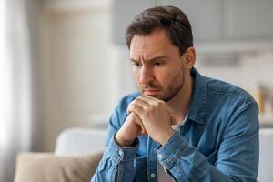 A man, dressed in a blue button-down shirt, sits on a couch, contemplating something, with a thoughtful expression on his face. His hands are clasped together, resting beneath his chin, copy space photo