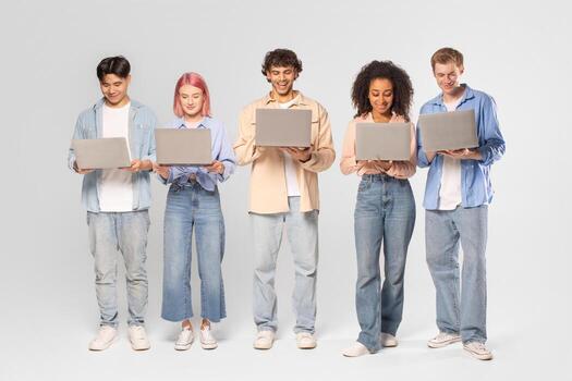 A diverse group of five young adults stand in a row, each using a laptop computer. They are all dressed casually and appear to be focused on their devices. The background is simple white photo