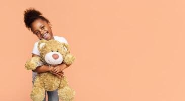 A little Black girl with curly hair smiles at the camera while holding a brown teddy bear against a peach-colored background, copy space photo