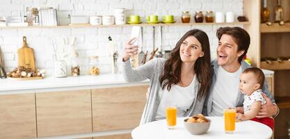 A happy family is taking a selfie in a modern kitchen. The woman is holding a smartphone and smiling at the camera, while the man holds their baby, copy space photo