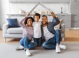 African American family seated on the floor facing a couch. They appear to be engaged in conversation or watching something together. Parents holding carton roof above their child photo
