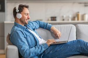 A man in a denim shirt sits on a light-colored couch, relaxed with headphones on and his laptop in his lap. He is smiling, looking at the screen of his computer. The room has a modern design photo