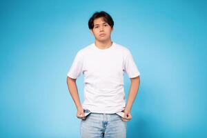 Asian young guy stands against a blue background, wearing a white shirt and jeans. He appears to be checking his pockets with a casual stance. photo