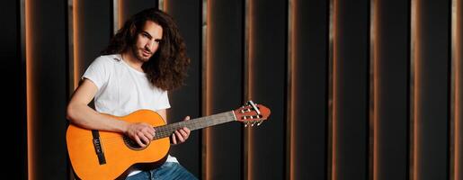 A young musician with long hair passionately strums an acoustic guitar in a modern studio setting. The warm light highlights his focused expression as he engages with his music, copy space photo
