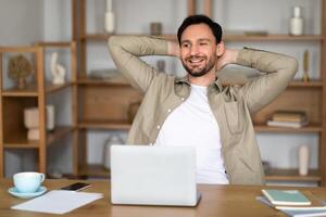 A young man with a cheerful expression sits back at his desk in a well-lit, contemporary office setting, hands clasped behind his head. In front of him is an open laptop, alongside a cup of coffee photo